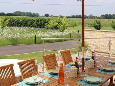 An outdoor dining table set with plates glasses and bottles on a patio overlooking a garden with trees and fields at The Anchorage @ Nables Farm in Upper Seagry