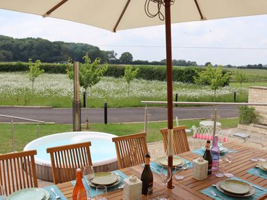 An outdoor dining area with a wooden table set with plates and glasses under an umbrella next to a round hot tub overlooking a field at The Anchorage @ Nables Farm in Upper Seagry