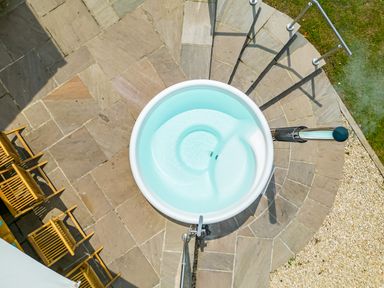 A round hot tub on a stone patio next to wooden chairs and a grassy area at The Anchorage @ Nables Farm in Upper Seagry
