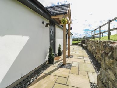 A paved pathway alongside a white building with a black door and potted plants near a wooden canopy and stone wall at Ty Cuddfan in Llangoed