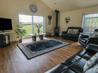 A living room with black leather sofas a rug a coffee table a wood stove and windows with vertical blinds at Ty Cuddfan in Llangoed