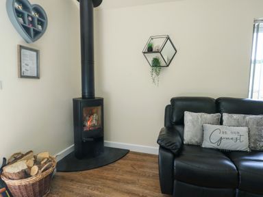 A corner with a black wood burning stove next to a basket of firewood and a black sofa with cushions at Ty Cuddfan in Llangoed