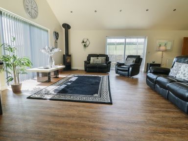 A living room with black leather sofas a black patterned rug a wood stove and large windows with vertical blinds at Ty Cuddfan in Llangoed