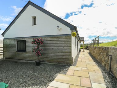 A paved pathway beside a house with wooden and white walls a potted flowering plant and a stone wall at Ty Cuddfan in Llangoed