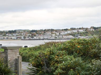 A view of a coastal town with houses on a hillside seen across water with bushes and a stone pillar in the foreground at Carriguisnagh in Ballycastle