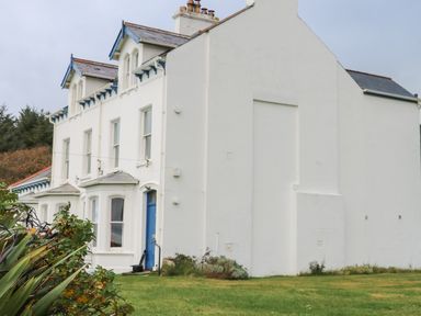 A white two-story house with blue trim and a blue door with plants and grass in the foreground at Carriguisnagh in Ballycastle