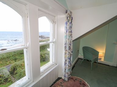 A room corner with a green chair next to a small lamp and floral curtain by windows overlooking the sea at Carriguisnagh in Ballycastle