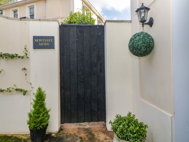 A black wooden gate in a white wall with a hanging lamp and plants at Monterey Mews in Torquay