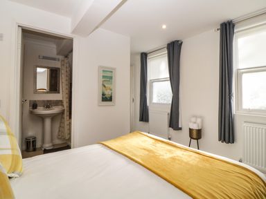 A bedroom with a bed covered with white and yellow bedding two windows with dark curtains and a bathroom visible through an open door at Monterey Mews in Torquay