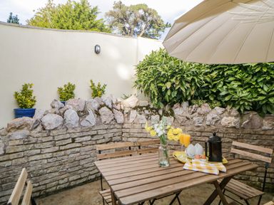 A wooden outdoor table with chairs a glass vase with flowers and breakfast items under a beige umbrella at Monterey Mews in Torquay