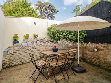 An outdoor patio area with a wooden table and six chairs under a large beige umbrella at Monterey Mews in Torquay