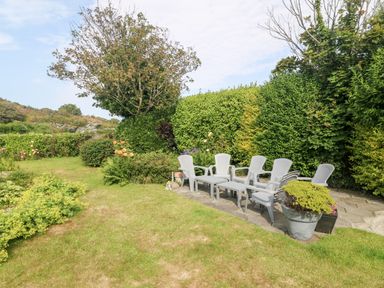 A garden with plastic chairs and tables on a stone patio surrounded by hedges and plants at Little Mead in Trearddur Bay