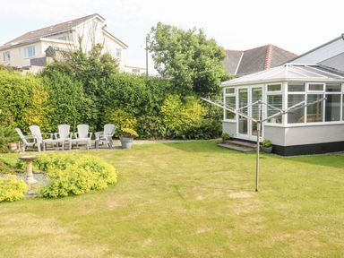 A backyard with a lawn, garden plants, a birdbath, plastic chairs around a table, and a conservatory at Little Mead in Trearddur Bay