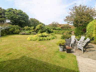 A garden with a grass lawn a flower bed plastic chairs on a patio and trees at Little Mead in Trearddur Bay