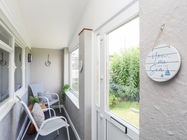 A narrow sunroom with two chairs a small wooden table potted plants and a round hanging sign next to a glass door at Little Mead in Trearddur Bay