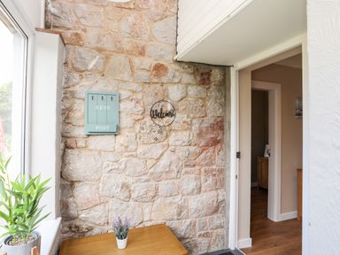 A stone wall with a key and post holder and a welcome sign next to a doorway with a wooden table holding a small plant near a window at Little Mead in Trearddur Bay