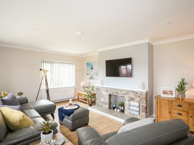 A living room with gray leather sofas a stone fireplace and a wall-mounted TV at Little Mead in Trearddur Bay
