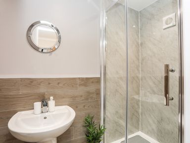 A bathroom with a pedestal sink a round mirror a shower with glass doors and a small plant at Little Mead in Trearddur Bay