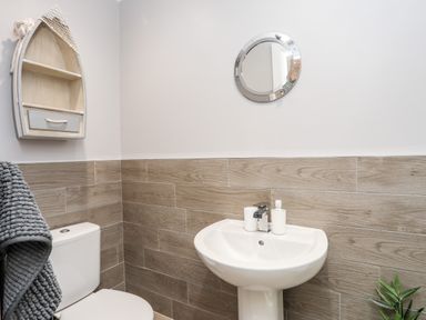 A bathroom with a toilet, pedestal sink with soap dispenser, round mirror, boat-shaped wall shelf, and a towel on a rack at Little Mead in Trearddur Bay