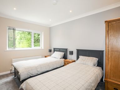 A bedroom with two single beds side by side with checked bedding two wooden nightstands with lamps a wooden wardrobe and a window with a view of greenery at Little Mead in Trearddur Bay
