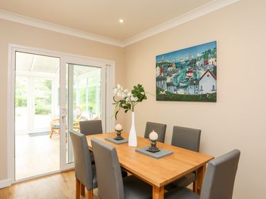 A dining room with a wooden table and six gray chairs with candles and a vase with flowers on the table and a painting on the wall at Little Mead in Trearddur Bay