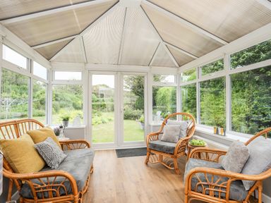 A conservatory with rattan chairs and sofa with cushions and a view of a garden at Little Mead in Trearddur Bay