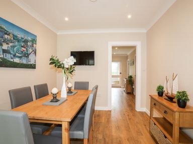 A dining room with a wooden table surrounded by six grey chairs a wall-mounted television a side table with plants and a model sailboat at Little Mead in Trearddur Bay