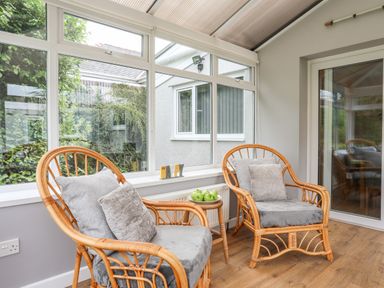 A sunroom with two wicker chairs with gray cushions and a small wooden table with green apples at Little Mead in Trearddur Bay