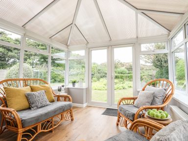 A conservatory with wicker furniture cushions a table with green apples and garden view at Little Mead in Trearddur Bay