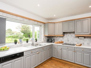 A kitchen with grey cabinets white countertops a double sink a microwave and a window overlooking a garden at Little Mead in Trearddur Bay