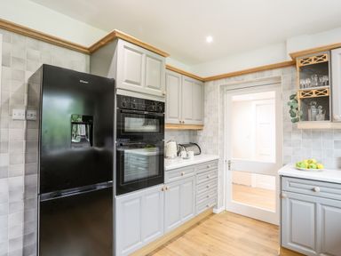 A kitchen with gray cabinets a black refrigerator double oven a kettle mugs a fruit bowl and a glass door at Little Mead in Trearddur Bay