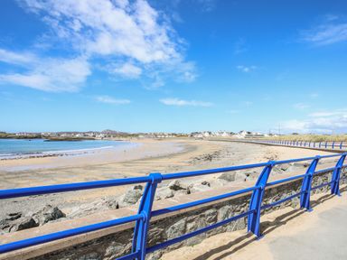 A sandy beach with a blue railing and rocky barrier overlooking a coastal town at Little Mead in Trearddur Bay