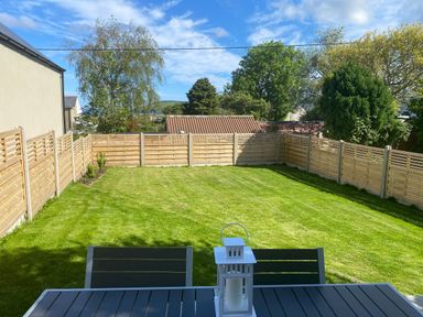 A fenced backyard with green grass and a patio table with chairs and a lantern on top at Awelfa in Newport Pembrokeshire