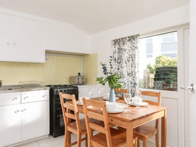 A kitchen with a wooden dining table set for four next to a window with patterned curtains and white cabinets at The Little Dolphins in Rhosneigr