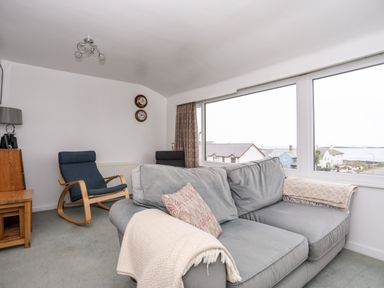 A living room with a gray sofa blue rocking chairs and large windows overlooking houses and the sea at The Little Dolphins in Rhosneigr