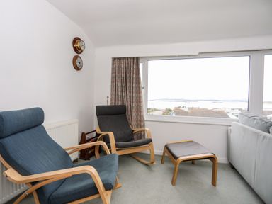A seating area with two armchairs and a footrest near a large window overlooking rooftops at The Little Dolphins in Rhosneigr
