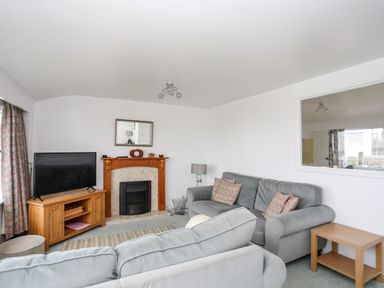 A living room with sofas a fireplace a television on a wooden stand and a window to another room at The Little Dolphins in Rhosneigr