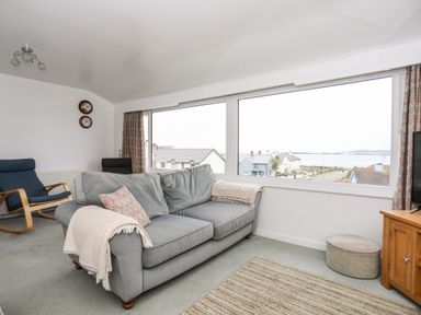 A living room with a grey sofa and large window overlooking houses and water at The Little Dolphins in Rhosneigr