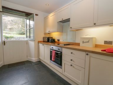 A kitchen with cream cabinets wooden countertops an oven toaster bread bin and a half door open to outside at Oak Tree Cottage in Coniston