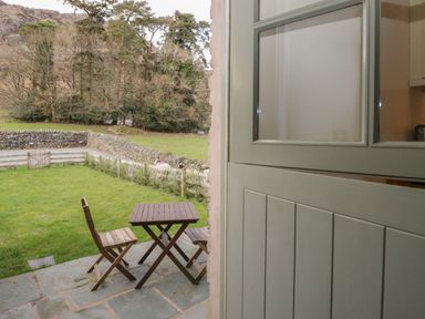 A wooden table and two chairs on a stone patio overlooking a green lawn with a stone wall and trees at Oak Tree Cottage in Coniston