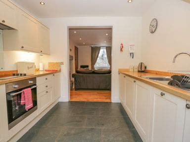 A kitchen with countertops on both sides an oven a sink and a view into a living room with a sofa and a window at Oak Tree Cottage in Coniston