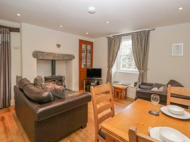 A living room with brown leather sofas a wood-burning stove a wooden dining table and chairs and a window with curtains at Oak Tree Cottage in Coniston