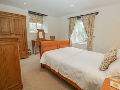 A bedroom with a wooden bed a wardrobe a dressing table a chair and two windows with patterned curtains at Oak Tree Cottage in Coniston