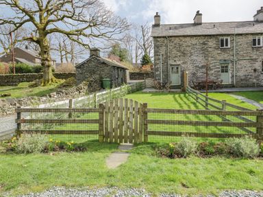 A fenced garden area with a wooden gate and stone houses in the background at Oak Tree Cottage in Coniston