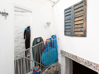 A small indoor space with a wetsuit hanging in a doorway a bodyboard and blue bags leaning against the wall a bucket with beach toys on the floor and rustic wooden shutters on the wall at The Sail Loft in St Ives