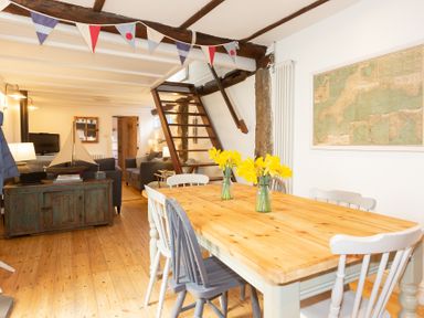 A dining area with a wooden table and chairs with flowers on the table and a living room with sofas and a TV in the background at The Sail Loft in St Ives