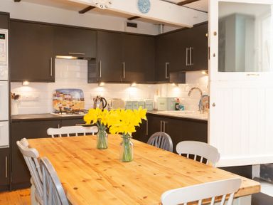 A kitchen with a wooden table and yellow flowers in jars with dark cabinets and white tiled backsplash at The Sail Loft in St Ives