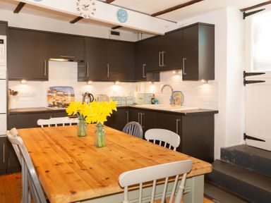A kitchen with dark cabinets white tiled backsplash wooden table with yellow flowers and mixed chairs at The Sail Loft in St Ives