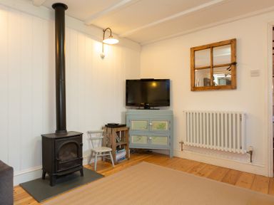 A room with a black wood stove a small blue cabinet with a TV on top a wooden crate with books a small chair a radiator and a wooden framed wall mirror at The Sail Loft in St Ives