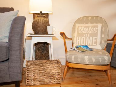 A chair with a cushion and a magazine next to a side table with a lamp and a cup at The Sail Loft in St Ives